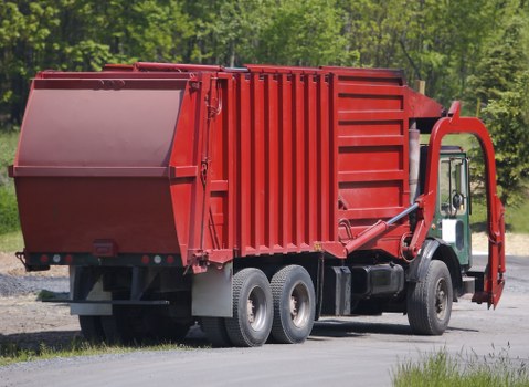 A large red industrial waste collection truck parked on a paved surface in a rural or semi-urban environment with a backdrop of green trees and grass. The truck features a prominent rectangular waste container with vertical ribbing along its sides, constructed from metal with a smooth painted finish. The container's rear section is open, designed for loading rubbish, and the front of the truck has a cabin with a visible door, side mirror, and window. The vehicle is equipped with six wheels, three on each side, with black rubber tires and silver rims. The truck's hydraulic lifting mechanism, with visible metal arms and joints, is positioned on the right side near the waste container, indicating its role in waste collection and disposal. The overall scene appears quiet and functional, emphasizing the practical purpose of facilitating rubbish removal services, typical of companies like Commercial Waste Removal Fitzrovia, serving areas potentially within London or nearby suburbs.
