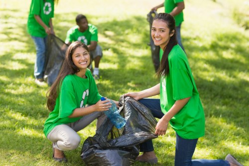 In an outdoor setting on a sunny day, four young individuals wearing green recycling-themed T-shirts participate in a community rubbish cleanup. Two women are at the forefront, smiling and kneeling on a grassy area with lush green trees in the background, actively gathering litter into large black trash bags. The woman on the left, with long brown hair, holds a water bottle in one hand and a filled trash bag, while the woman on the right, with long dark hair, is gripping an opened black bin bag ready to receive rubbish. Behind them, two more volunteers are visible; one is crouching and placing items into another trash bag, and the other stands holding a black bag, both contributing to the cleanup effort. The scene captures a collaborative, environmentally conscious activity focused on waste collection, with the volunteers wearing bright green T-shirts symbolizing recycling or environmental awareness. This image illustrates community engagement in waste management, highlighting the importance of local rubbish removal initiatives, potentially associated with companies like Commercial Waste Removal Fitzrovia, operating in the London area.