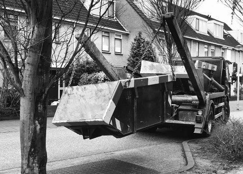 A black tow truck equipped with a hydraulic lift is parked on a residential street in Fitzrovia, London, during daytime. The truck's flatbed is raised and extended outward at an angle, with a large, rectangular metal container or storage box secured on it, showing signs of wear with dirt and scratches. The vehicle is positioned near the curb, with the front of the truck on the right side of the image and the rear extending towards the left. In the background, there are row houses with pitched roofs, brick facades, and sash windows, typical of urban residential areas. Leafless trees line the street, indicating a late autumn or winter season, and the pavement is visible beneath the truck. The environment appears quiet and typical for a city neighborhood, subtly indicating a context suitable for rubbish collection or clearance services provided by Commercial Waste Removal Fitzrovia, aligned with local waste management needs.