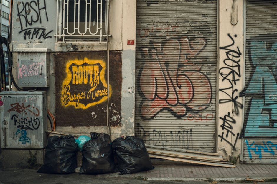 In front of a commercial building with graffiti-covered walls and metal shutter doors, five large black rubbish bags are placed on the pavement, filled with waste and tied at the top. Adjacent electri
