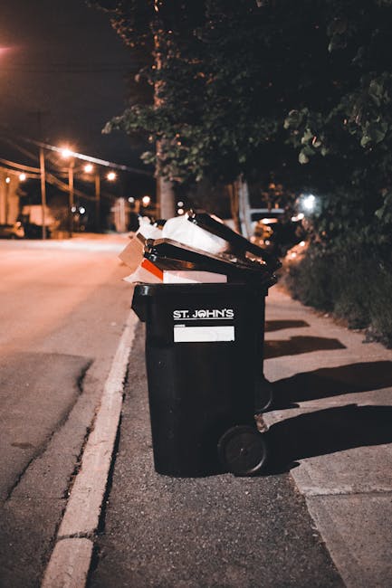A large green wheeled waste container filled with bags of garden or building waste materials, placed outdoors on a concrete pavement beside a dark painted brick wall. The bags are red with white text 