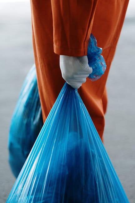 A male worker wearing a high-visibility vest with reflective orange and black trousers, a blue cap, and gloves is operating a leaf blower or vacuum device to clear litter from a grassy park area. The