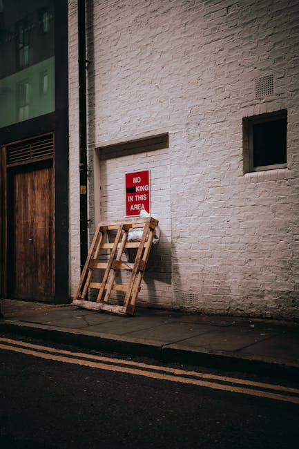 A collection of black garbage bags, some torn open revealing mixed waste, is piled directly in front of a worn, red metal fire exit door on an exterior wall. The bags are stacked haphazardly, with som