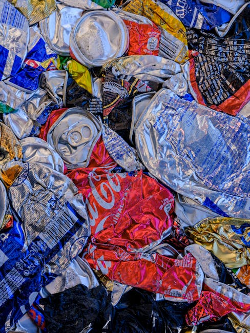 A close-up view of a pile of crushed aluminum beverage cans, predominantly silver with some red, blue, and black branding visible, including a partially readable Coca-Cola logo on one can. The cans ar